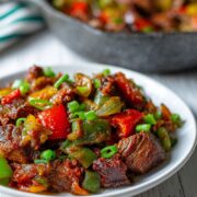 A plate of Asun, spicy roasted goat meat garnished with green peppers, red chili, and onions, served hot and smoky.