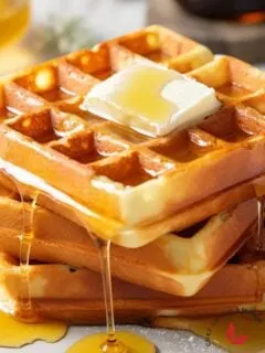 A stack of golden-brown classic waffles topped with melted butter and drizzled with maple syrup, ready to be served.