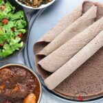 Injera in a tray with doro wat stew, salad, and Gomen wat served in bowls